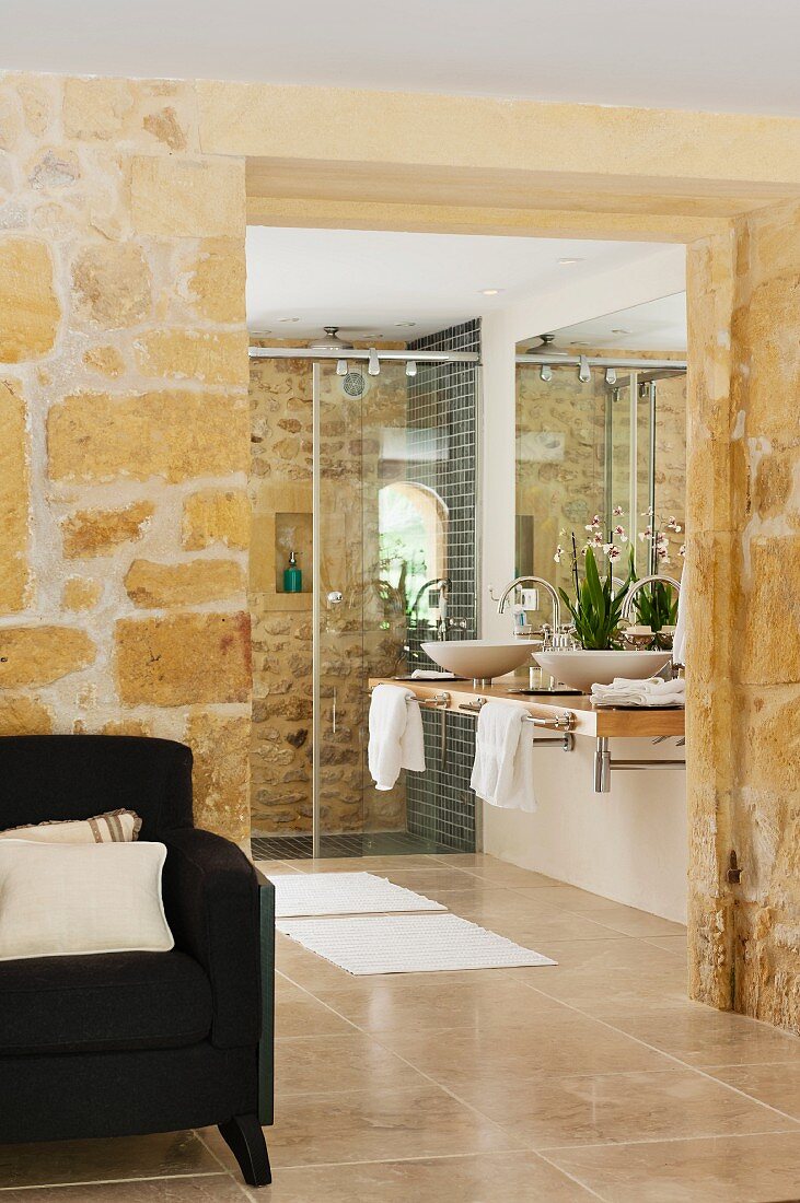 Open doorway in stone wall with view of washstand and shower in modern bathroom; armchair in foreground
