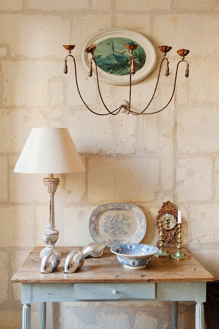 Nostalgic, shabby-chic table with drawer decorated with table lamp, antique crockery and ornamental shells against limestone wall