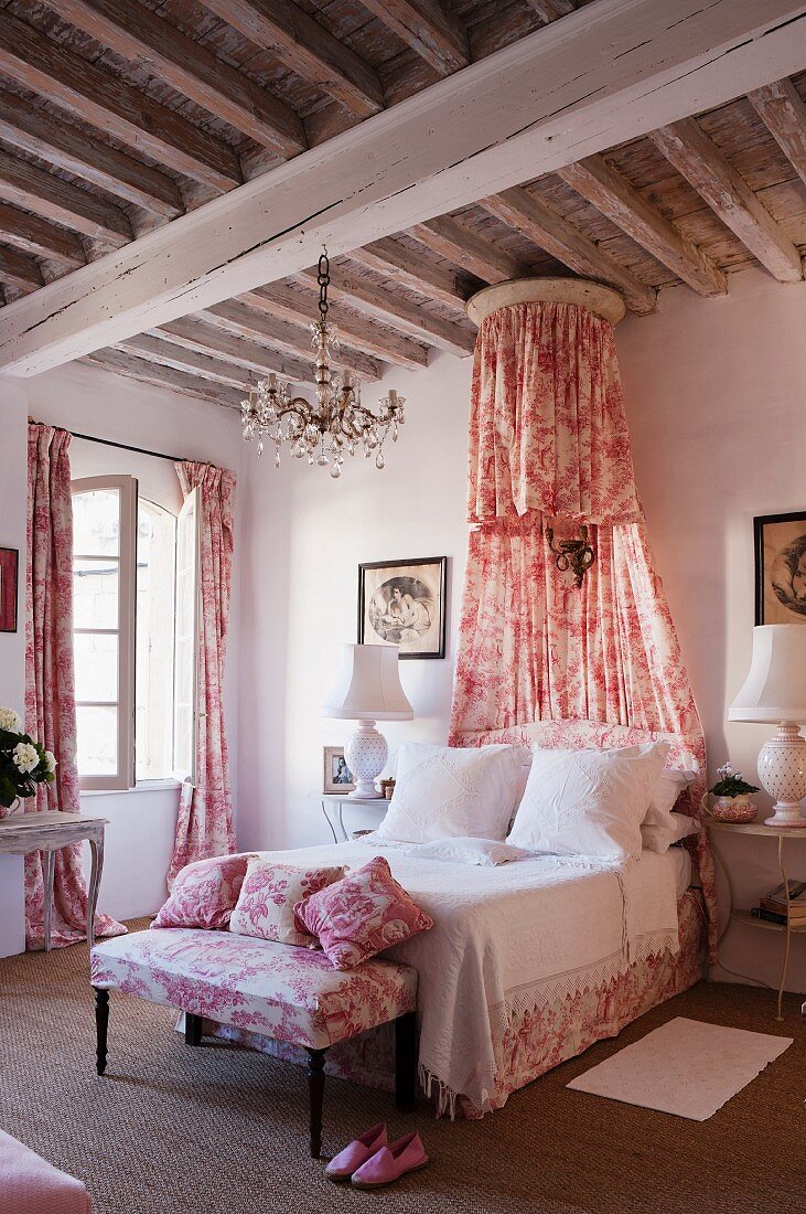 Romantisches Schlafzimmer mit rotweißem Toile-de-Jouy für Vorhänge, Kleiderbank, Kissen und Betthimmel im Shabby-Chic Flair unter rustikaler Holzbalkendecke
