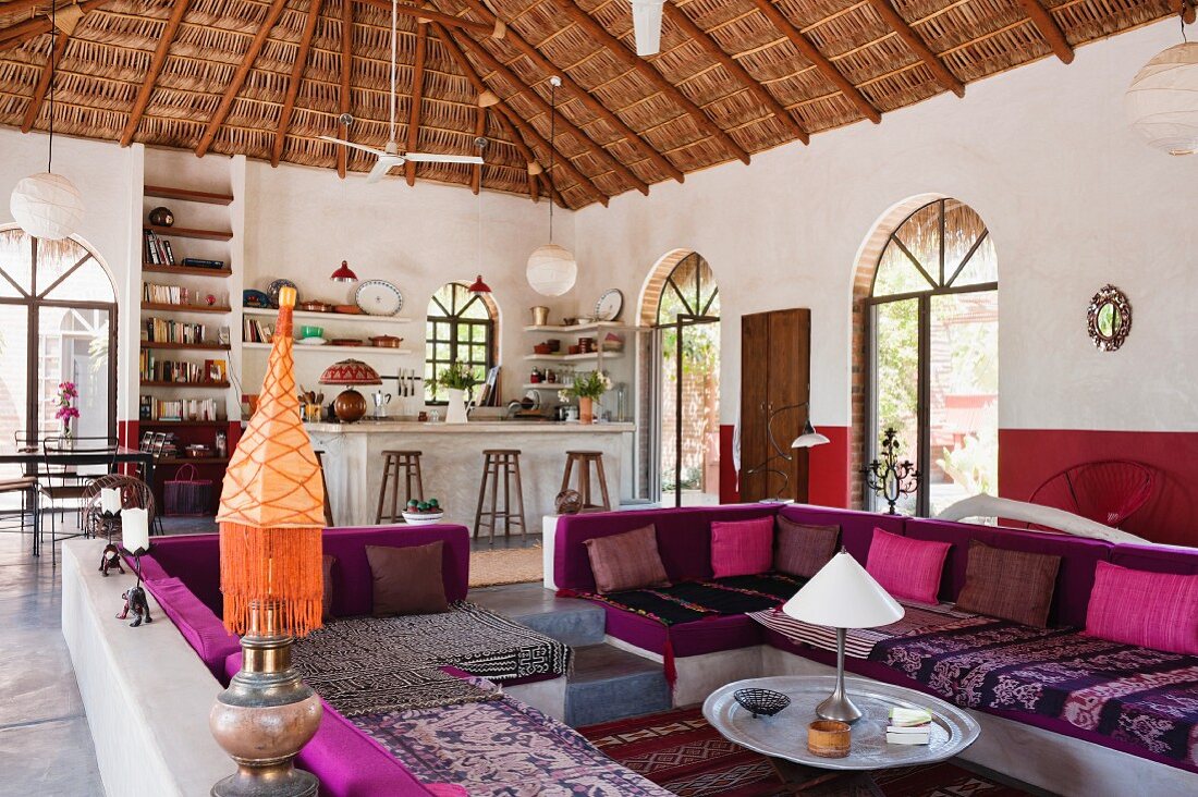 Lounge area with colourful seat cushions on masonry benches in open-plan interior with palm thatch roof
