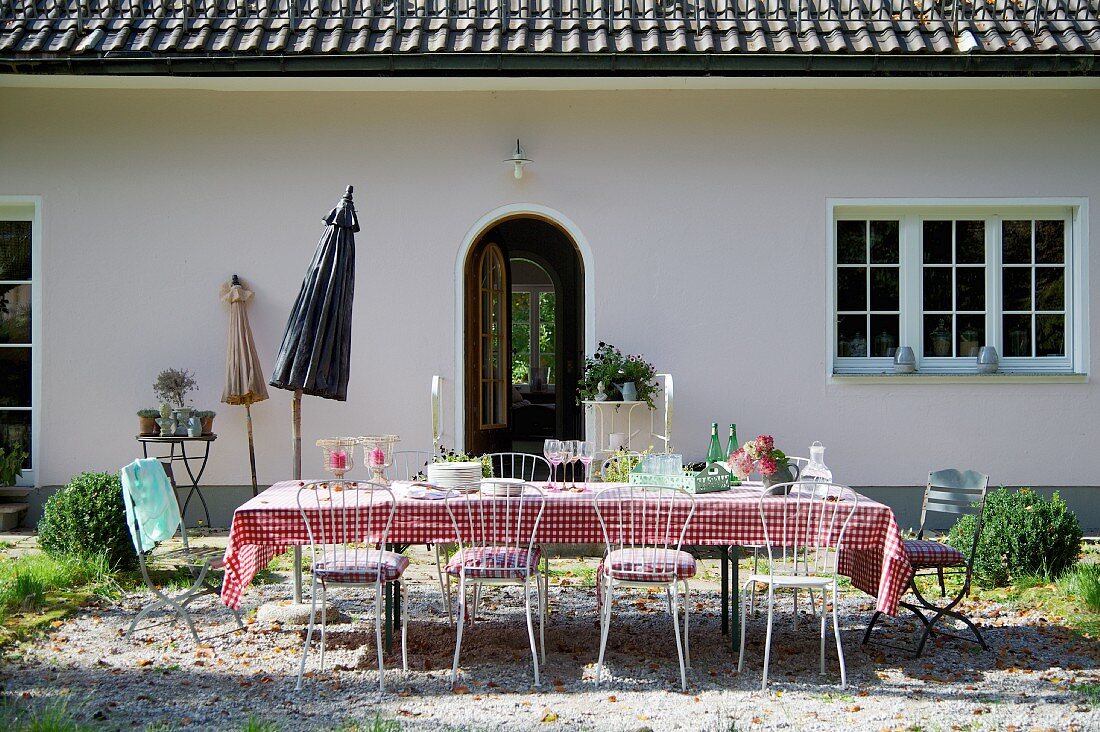 Gedeckter Tisch und filigrane Metallstühle auf gekiestem Platz im Garten, vor Wohnhaus in ländlichem Stil