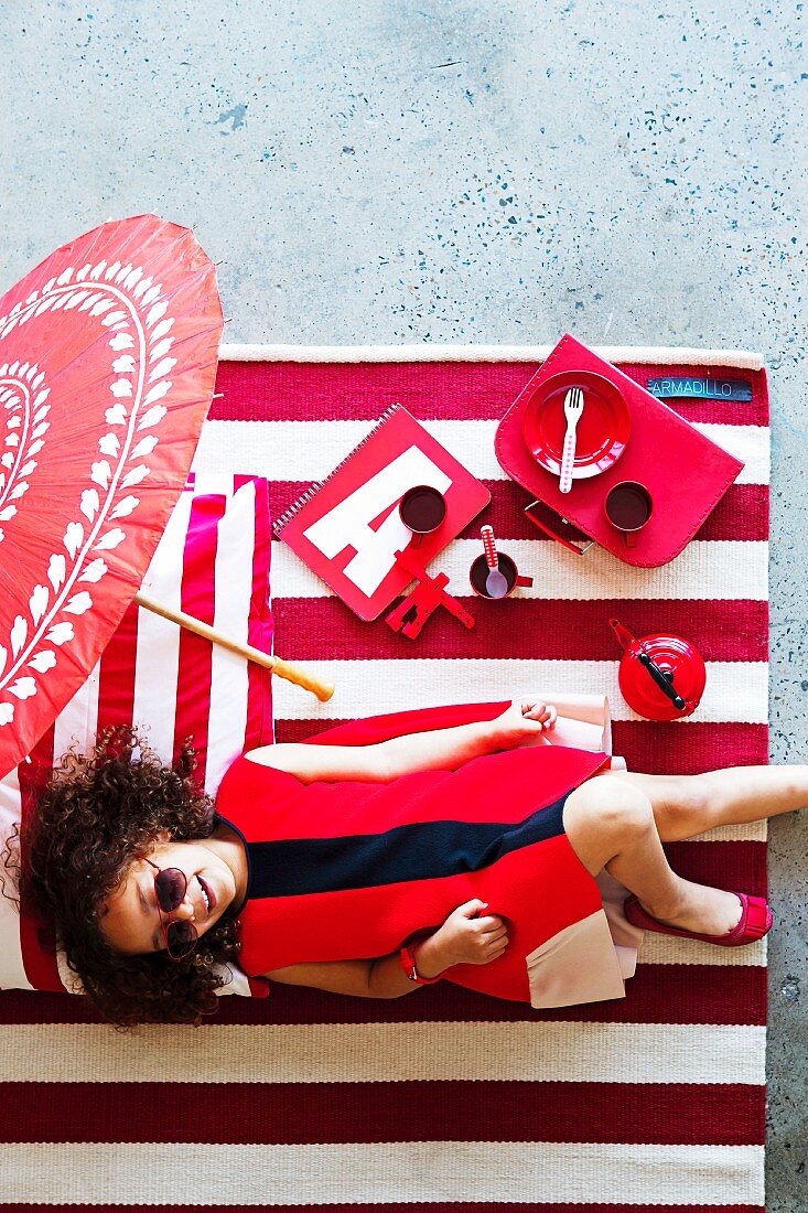 Rote Mädchenaccessoires und Spielsachen auf rot-weiss gestreiftem Teppich; darauf ein lachendes Mädchen im roten Kleid