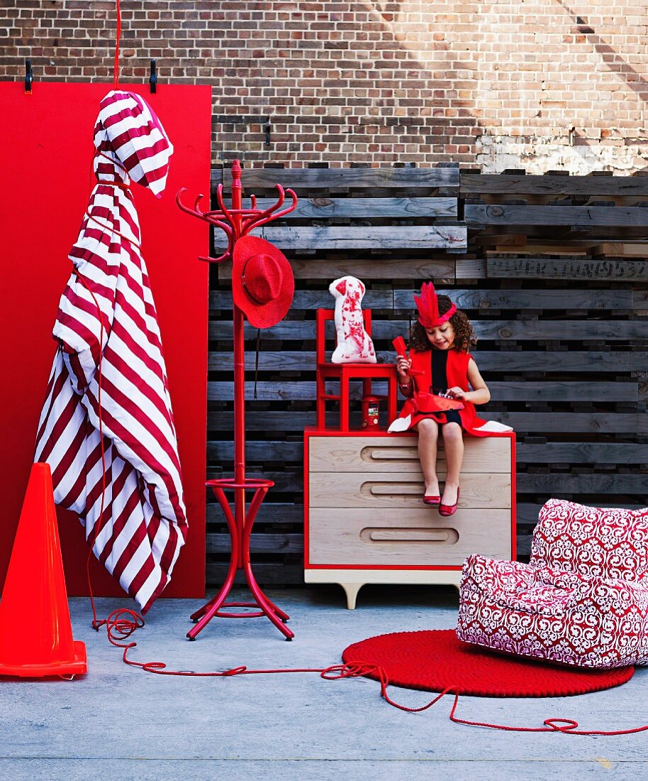 Wohnaccessoires und Möbel in kräftigem Rot; dazwischen ein Mädchen im roten Kleid auf einer Kommode sitzend