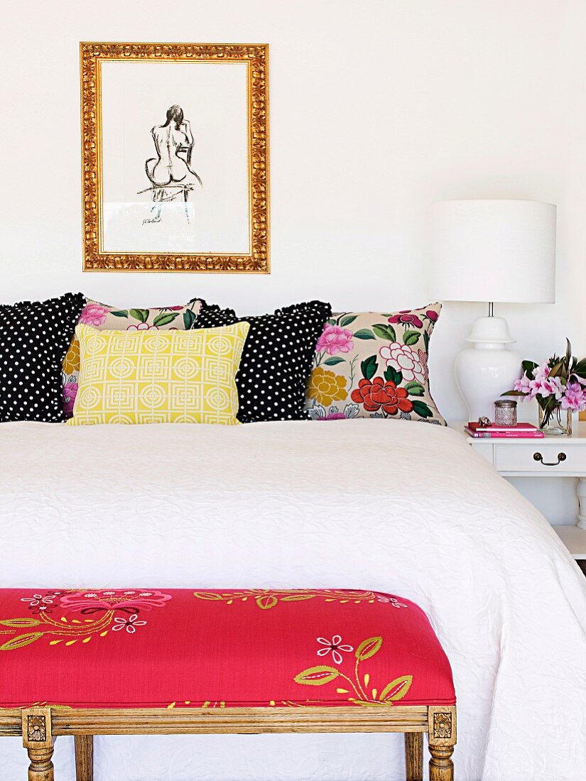 Double bed with white throw and several decorative pillows in different patterns; at the foot end a red upholstered stool with a floral motif