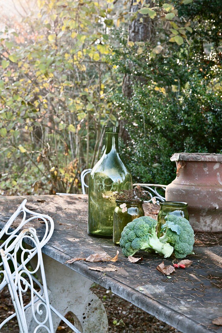 Herbstliches Stillleben im Garten mit Brokkoli, Gläsern und Amphore auf Steintisch
