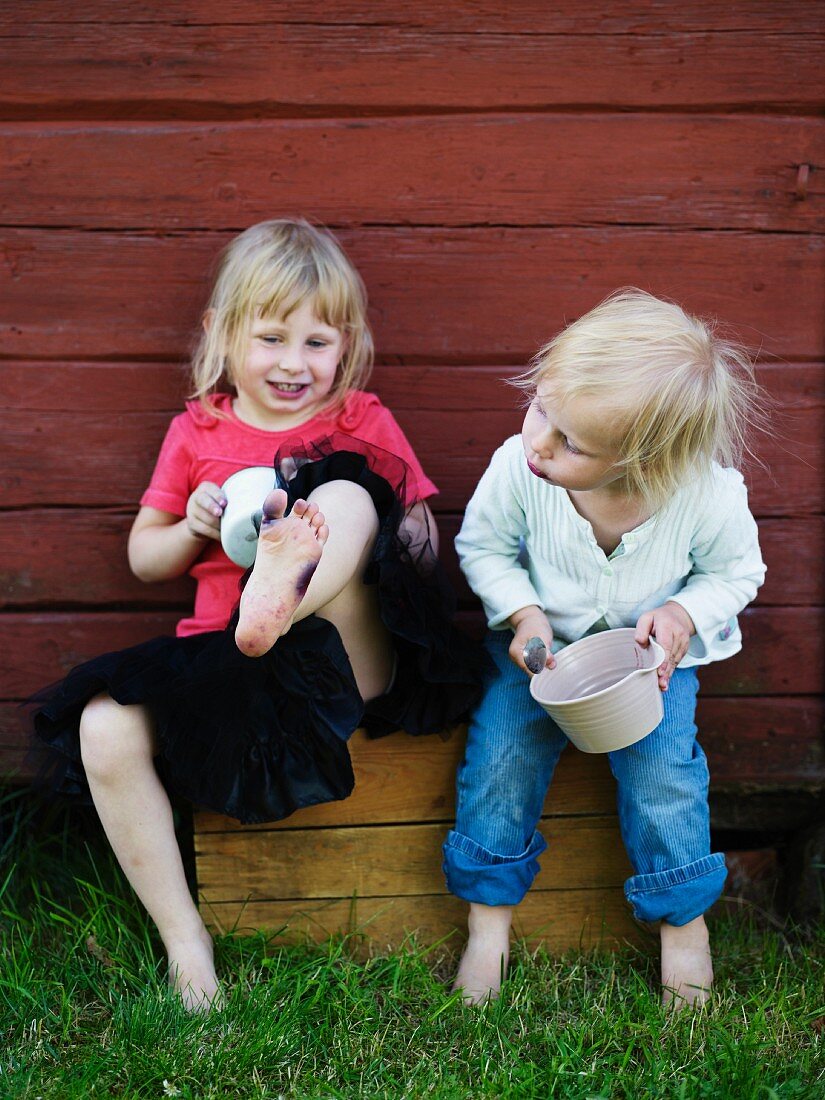 Mädchen mit Töpfen in der Hand sitzen und spielen vor Holzhauswand