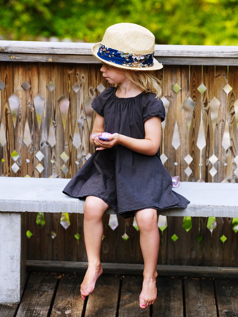 Mädchen mit Strohhut und Spielzeug-Telefon sitzt auf einer Bank