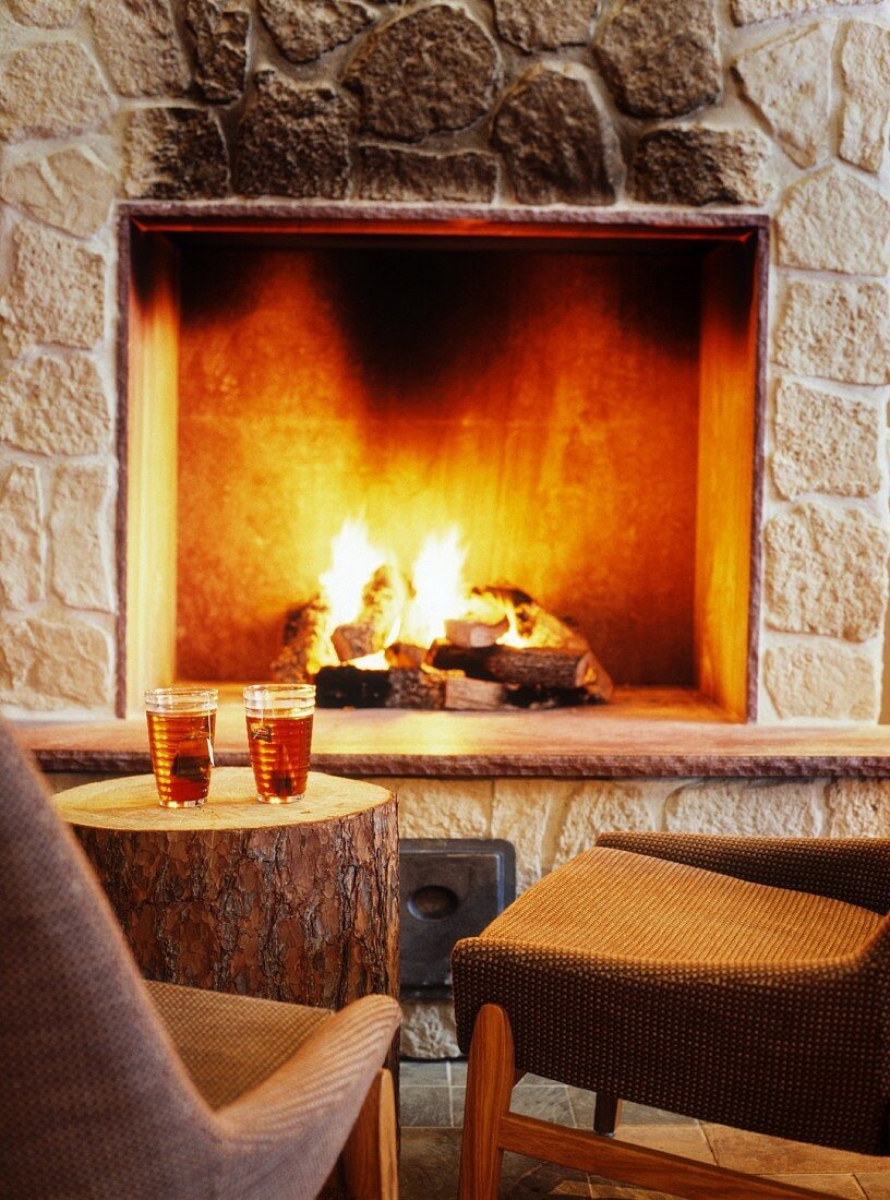 Warm drinks on tree stump used as side table and upholstered chairs in front of open fireplace