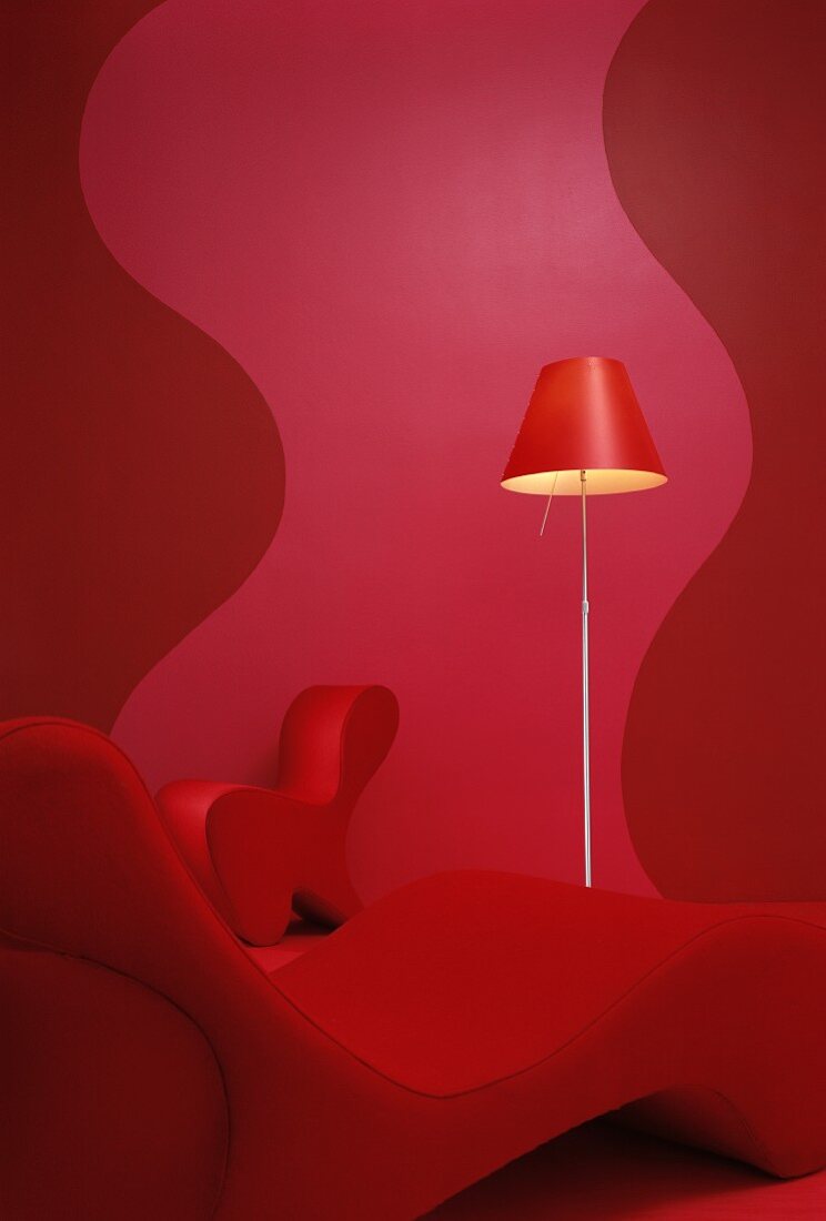 Red, curved lounger in front of standard lamp and wall with wavy pattern in different shades of red