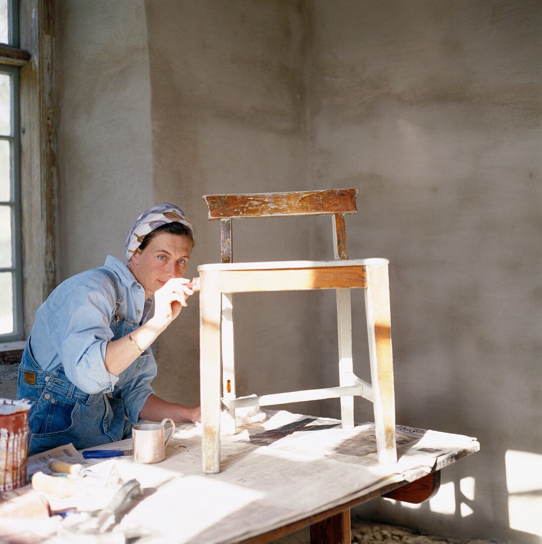 Woman in overalls painting wooden chair on table covered with newspaper