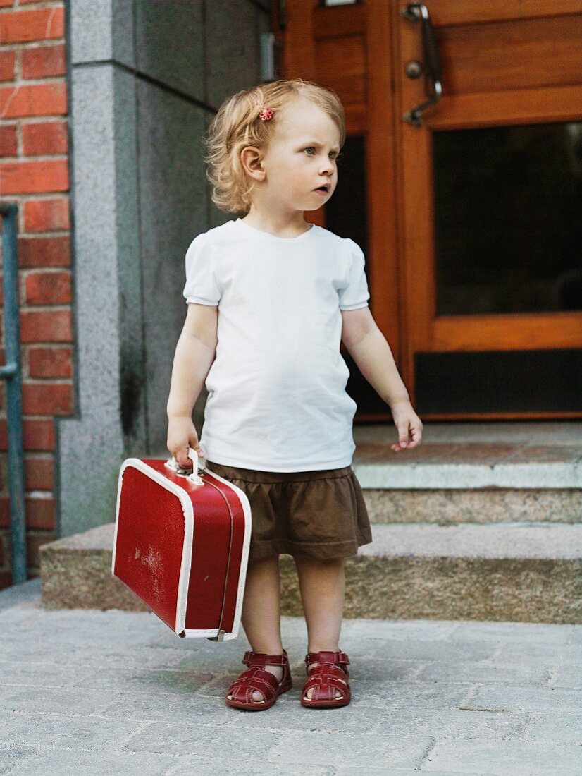 Blondes kleines Mädchen, mit rotem Koffer in der Hand, wartend vor einer Haustür