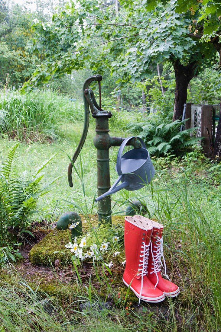 Watering can hanging from old hand pump in overgrown garden behind red wellingtons
