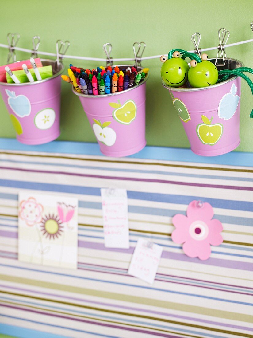Pens in metal beakers with printed patterns pegged on cord above striped pinboard with pictures of flowers