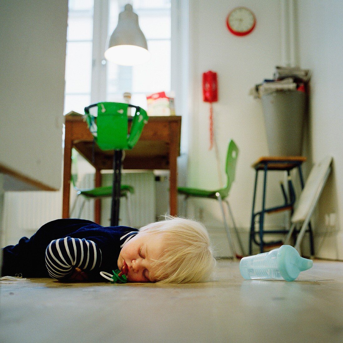Kind mit herausgefallenem Schnuller, eingeschlafen auf dem Küchenboden; dahinter knallgrüner Kinderhängestuhl am Esstisch