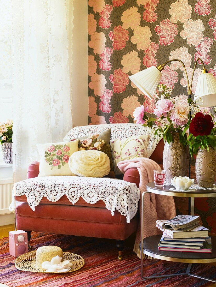 Upholstered armchair overloaded with lace cloths and rose-patterned cushions in front of wallpaper with large floral motif
