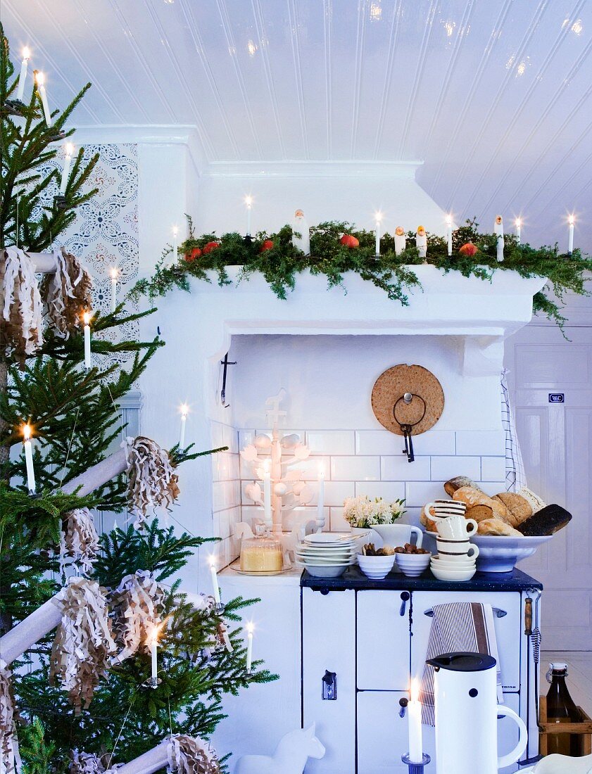 Geschmückter Weihnachtsbaum mit brennenden Kerzen in Landhausküche und Geschirrstapel auf Herd
