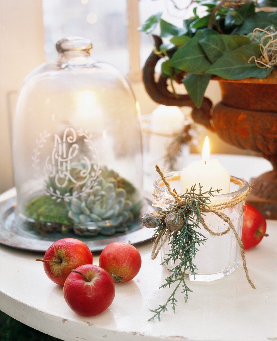 Lit candle with Christmas decorations next to apples and plants in various containers