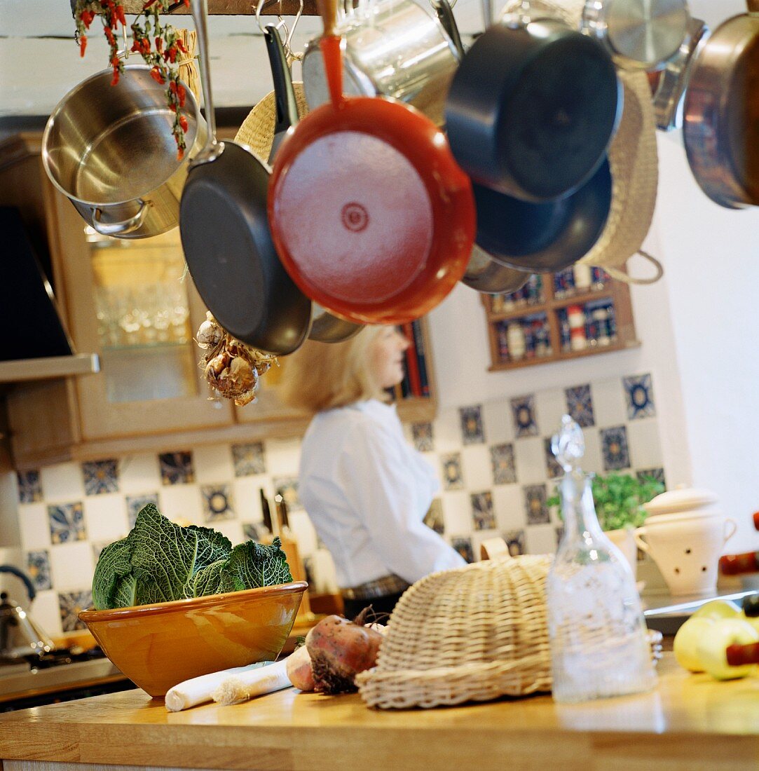 Von der Decke abgehängtes Kochgeschirr über Arbeitsplatte mit Gemüse und Blick auf Frau im Hintergrund