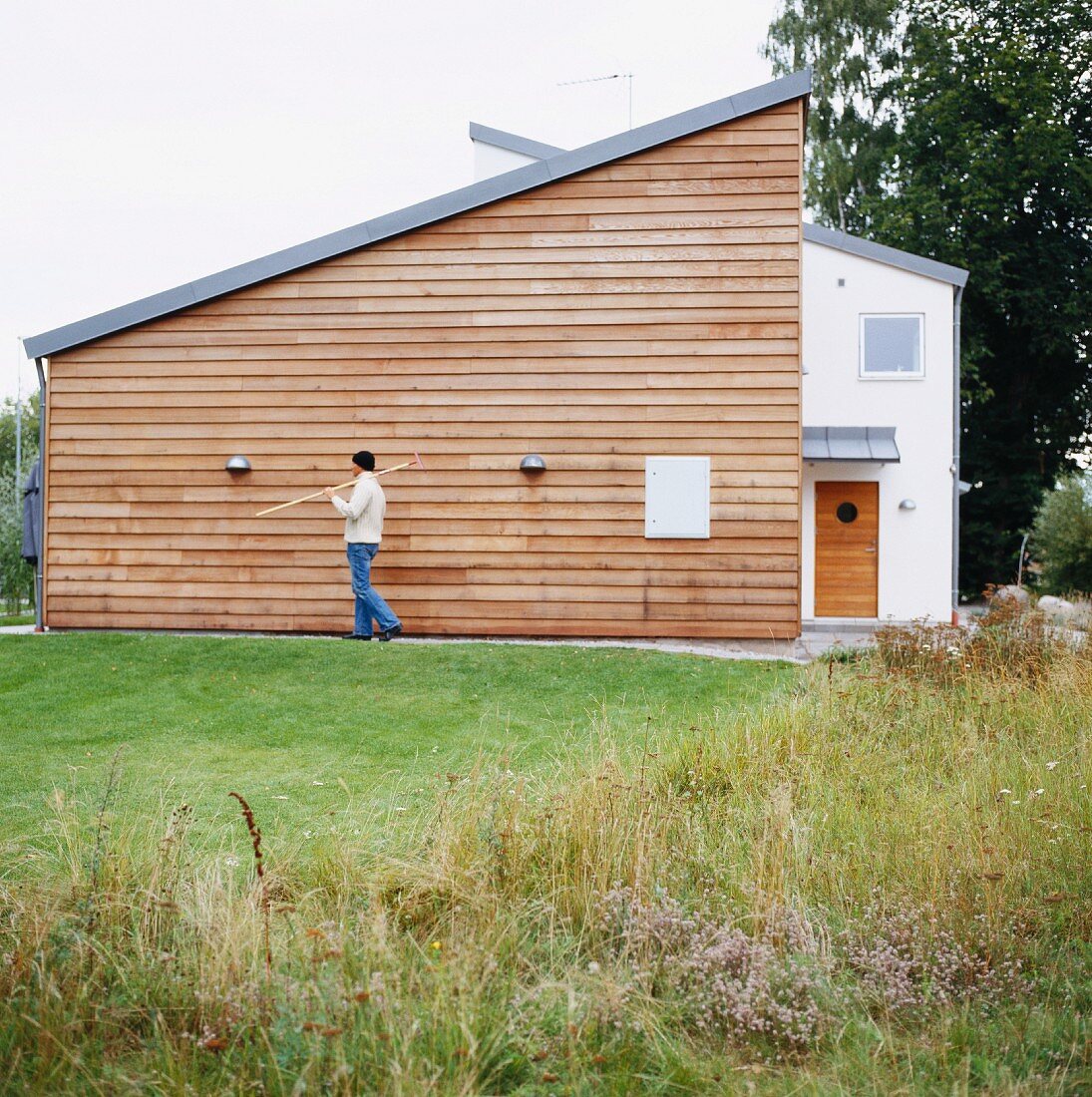 Rasenfläche vor modernem Wohnhaus und Mann mit Gartengerät vor Holzfassade