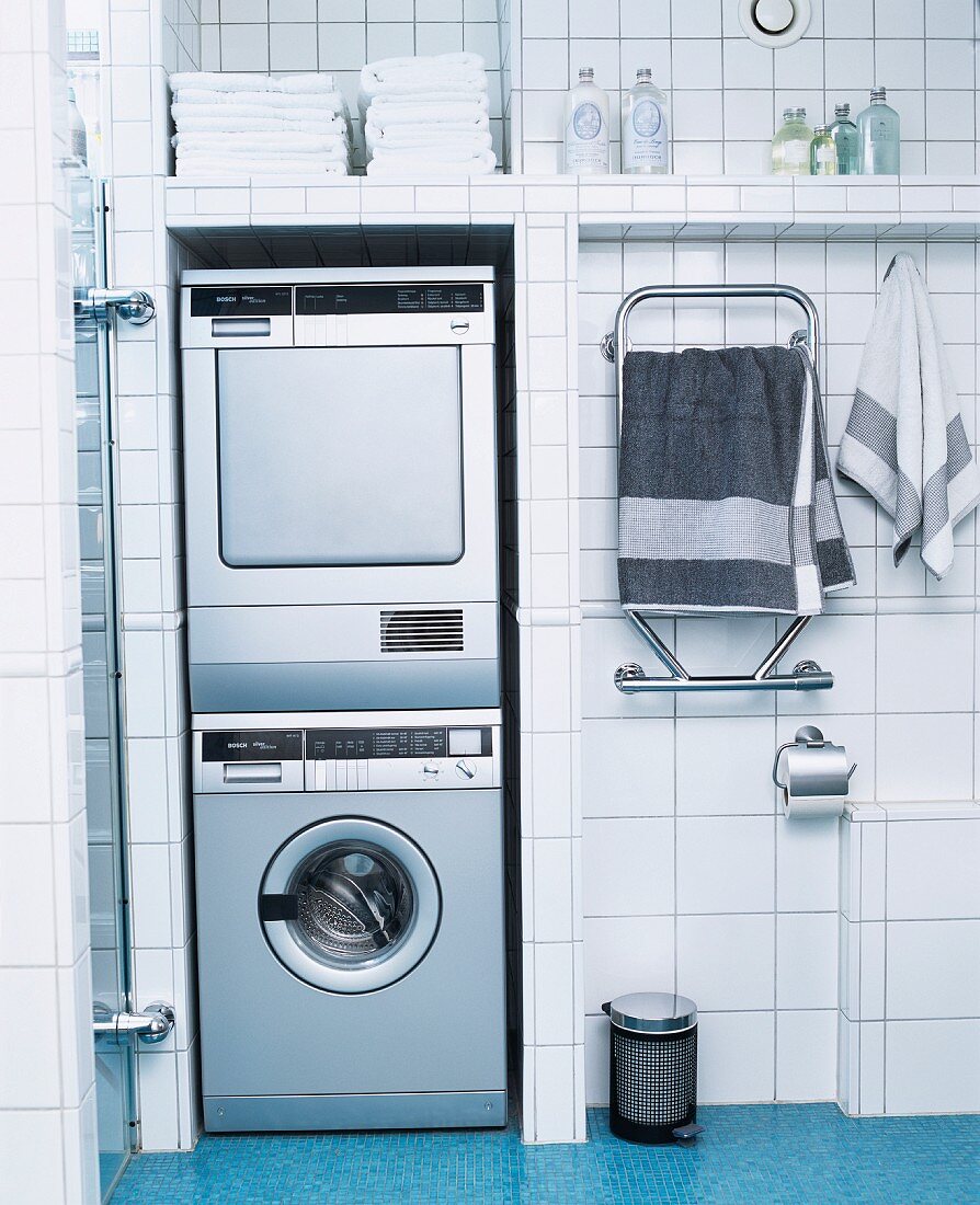 Waschmaschine und Trockner übereinander in Nische eines Badezimmers mit weissen Fliesen an Wand und blauen Mosaikfliesen an Boden