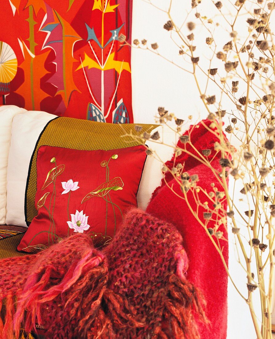Rote Strickdecke und Kissen auf Sofa neben Deko Bäumchen vor Wand mit rot gemusterter Tapete