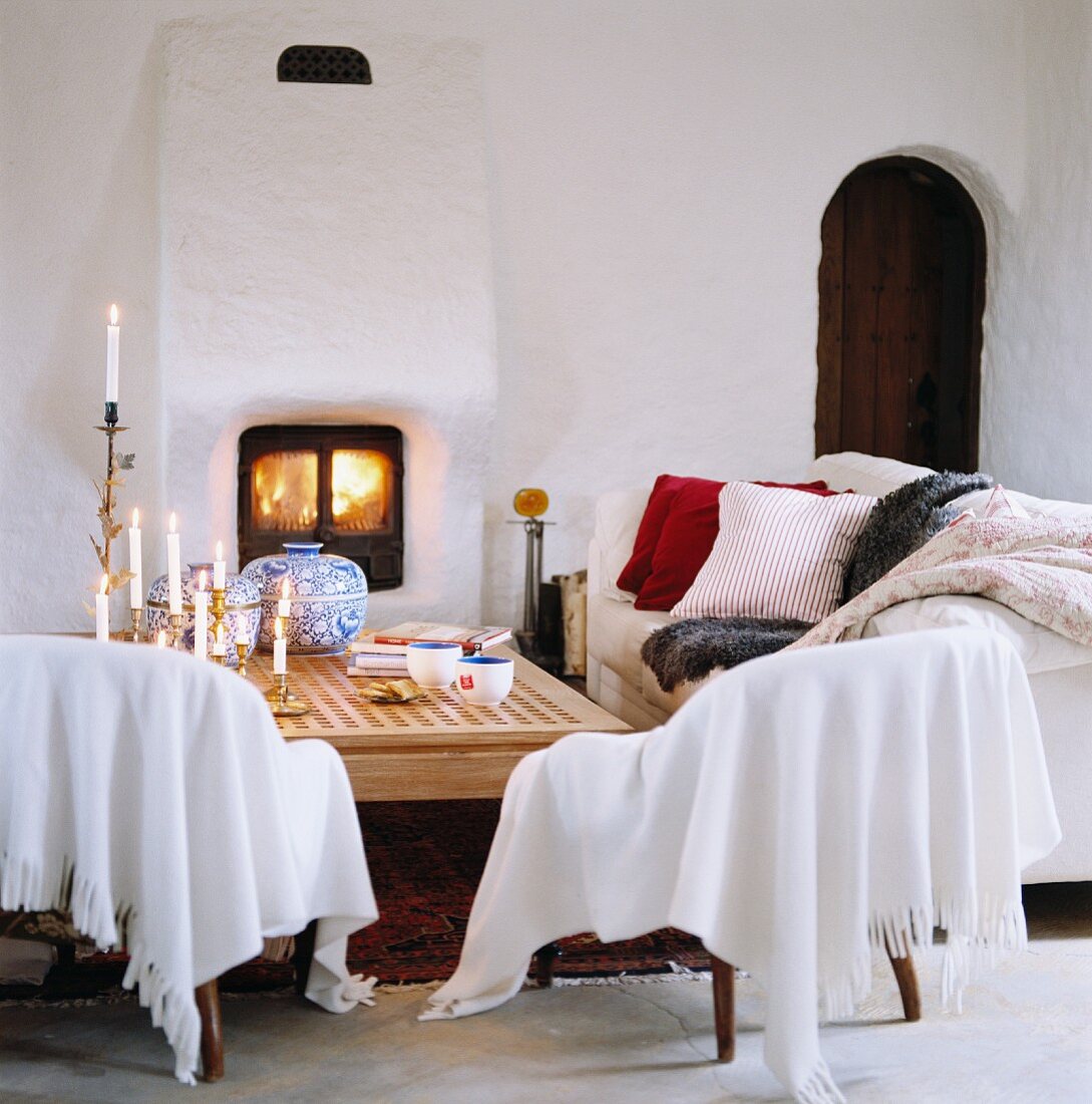 Decken und Kissen auf Sitzmöbeln vor gemauertem Kaminofen und rustikaler Rundbogentür