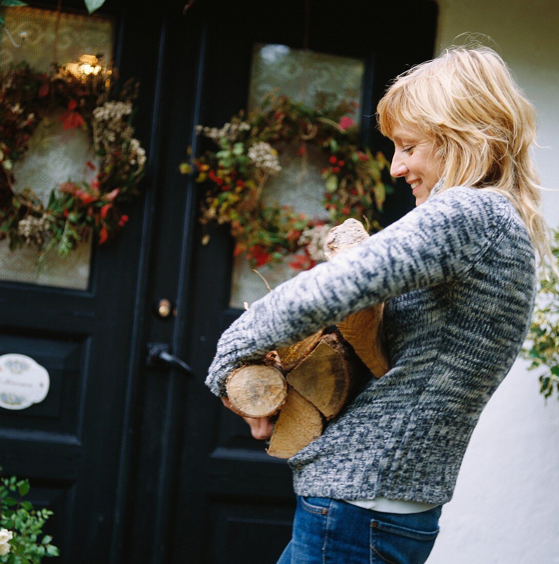 Junge Frau mit Holzscheiten vor Haustür im Landhausstil mit herbstlichen Dekokränzen