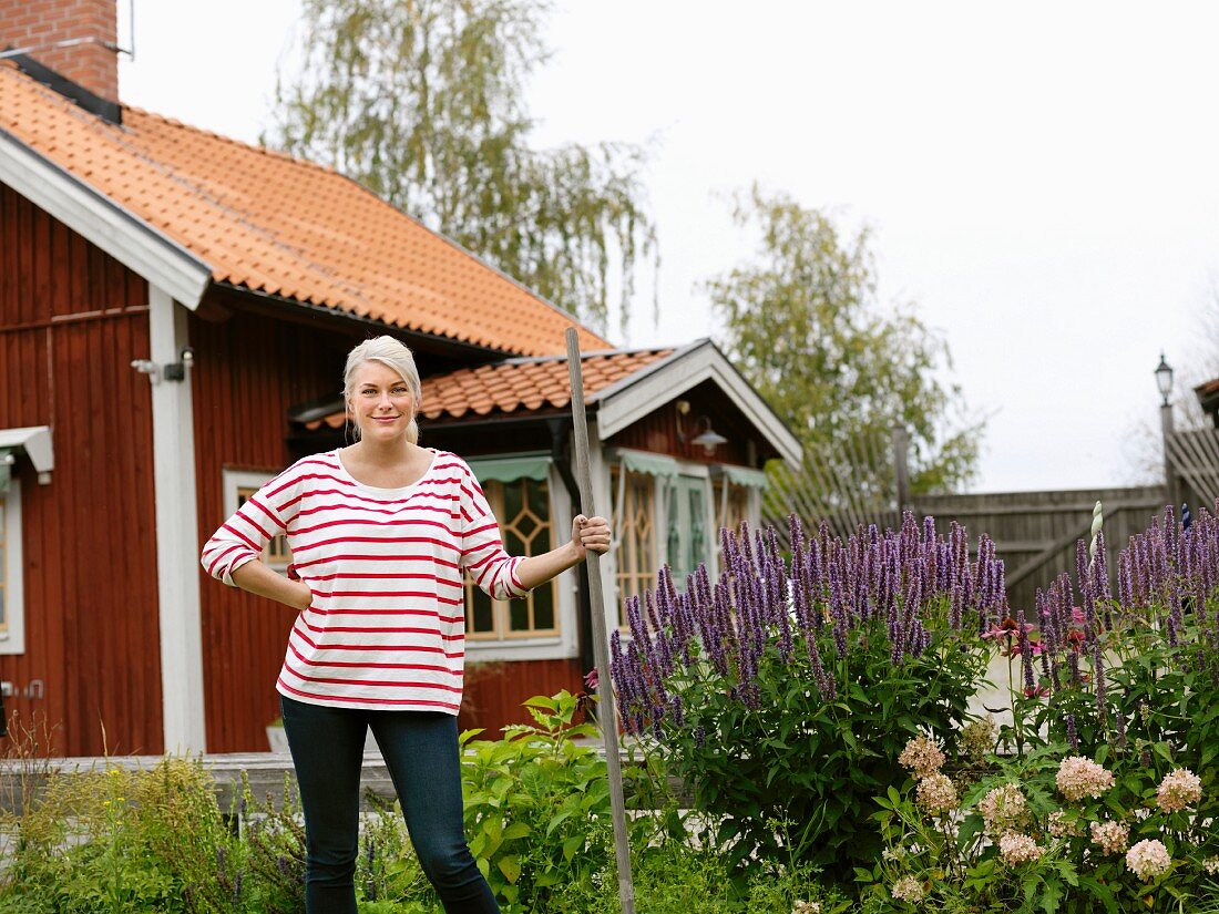 Junge Frau bei Gartenarbeiten neben blühendem Beet vor rotbraunem Holzhaus