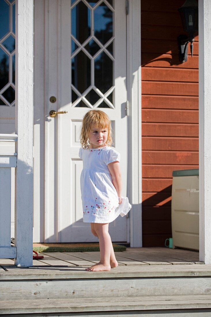 Kleines Mädchen in weißem Sommerkleid steht vor Haustür