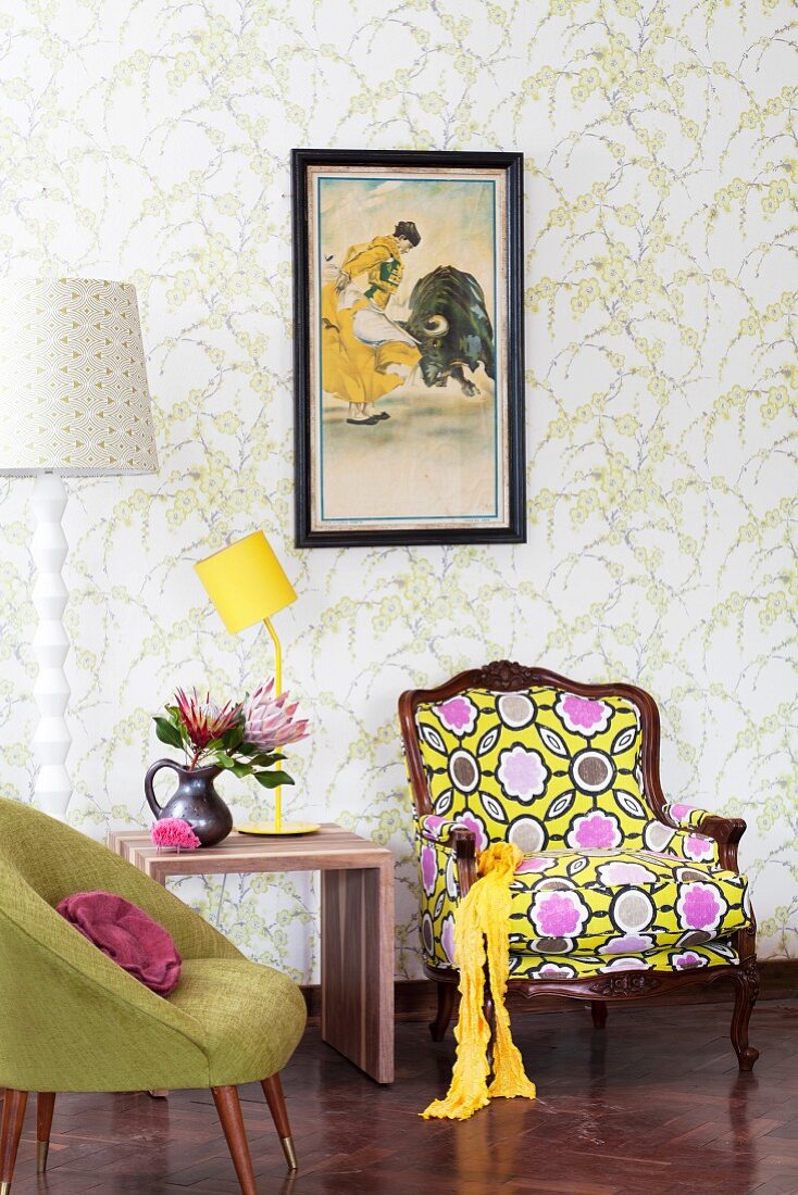 Fifties-Möbel neben peppigen, postmodernen Sessel und gelber Tischlampe auf Hocker vor Wand mit Blumenmotiven auf Tapete