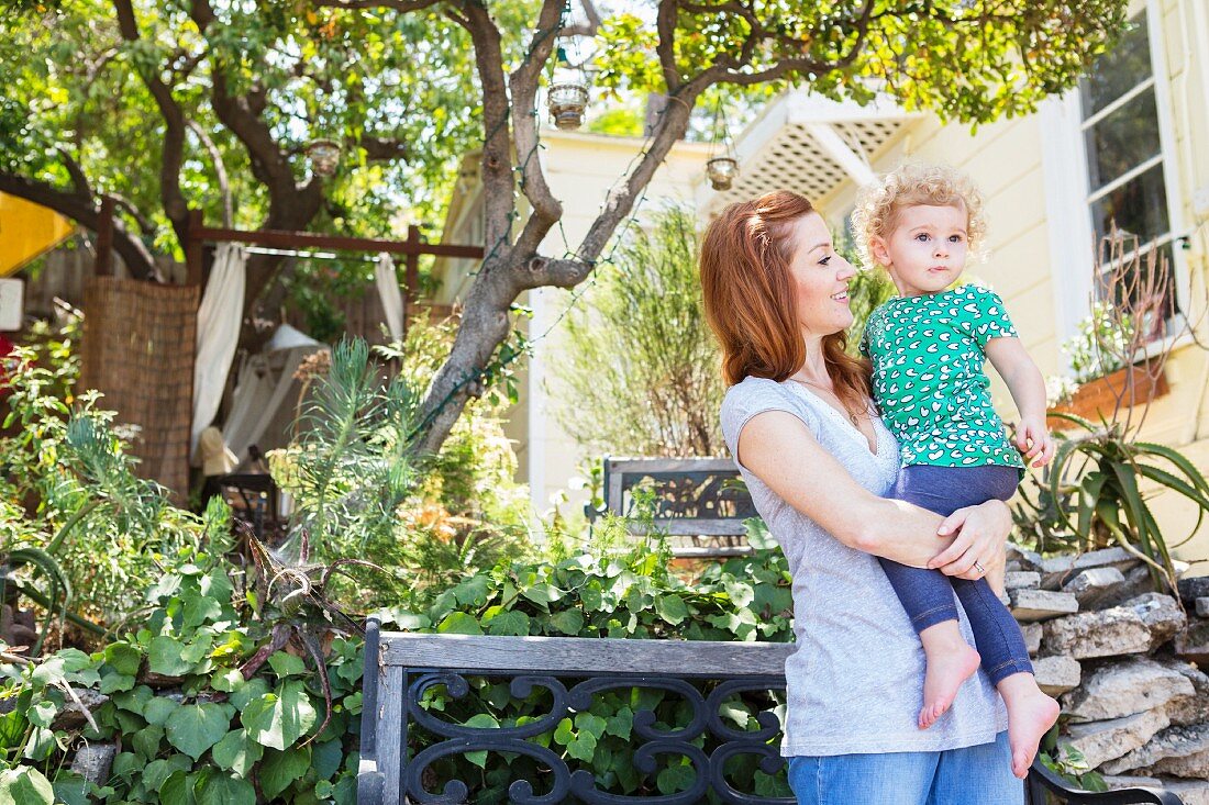 Junge Mutter mit Tochter auf dem Arm in Garten vor Holzhaus