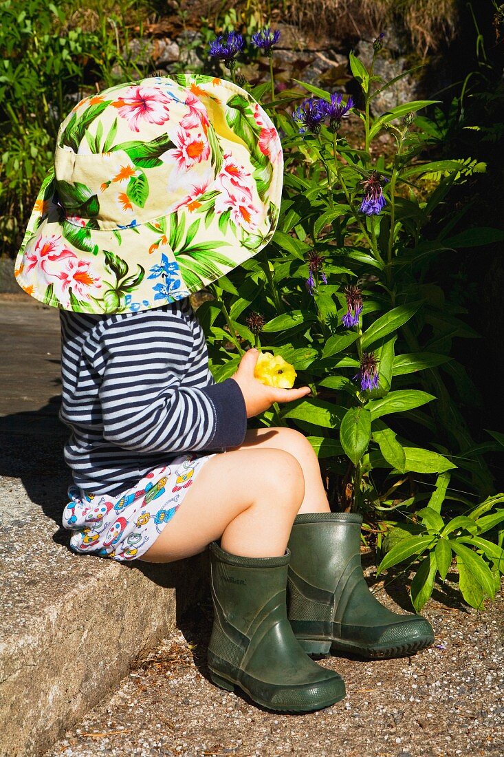 Kleines Kind mit Hut und Gummistiefel auf Steinstufe im Garten sitzend