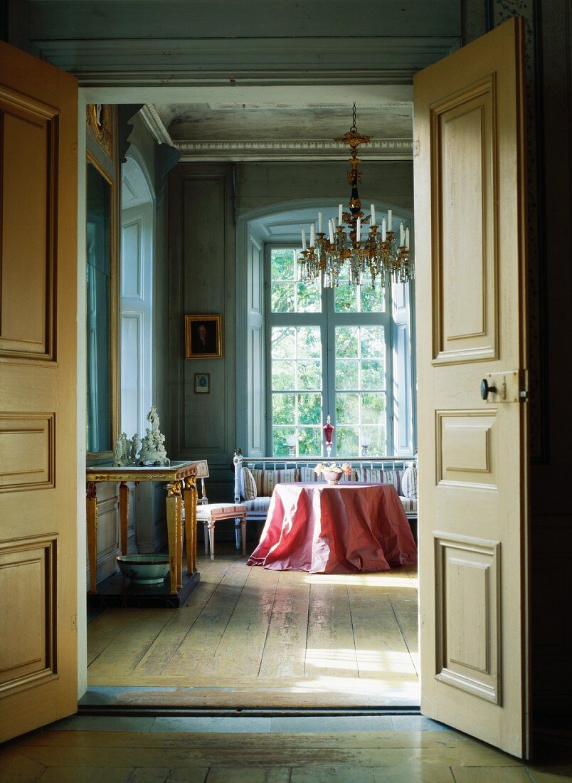 Offene Flügeltür und Blick auf Tisch mit rosa Tischdecke und Sitzbank an hohem Fenster im Wohnzimmer eines Schlosses
