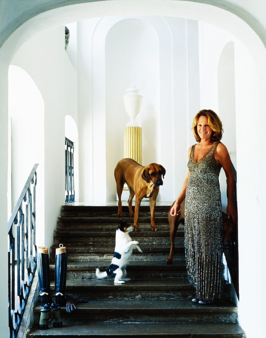 Frau im Abendkleid mit Hunden auf Treppe vor Nischen mit Rundbögen in herrrschaftlichem Treppenhaus
