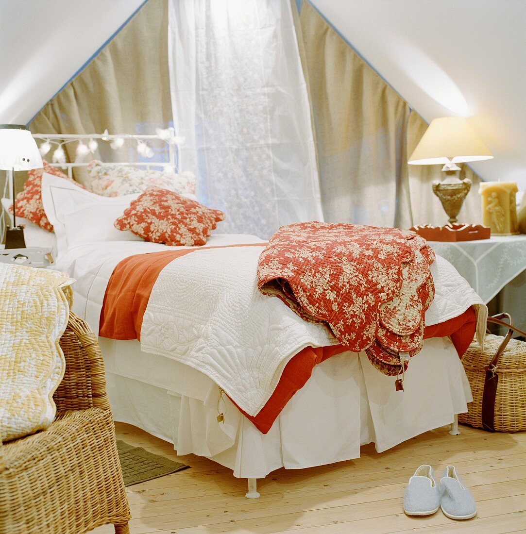 Bett mit verschiedenen Tagesdecken vor geschlossenem Giebelfenster zwischen Nachttischleuchten auf Tisch in romantischem Ambiente