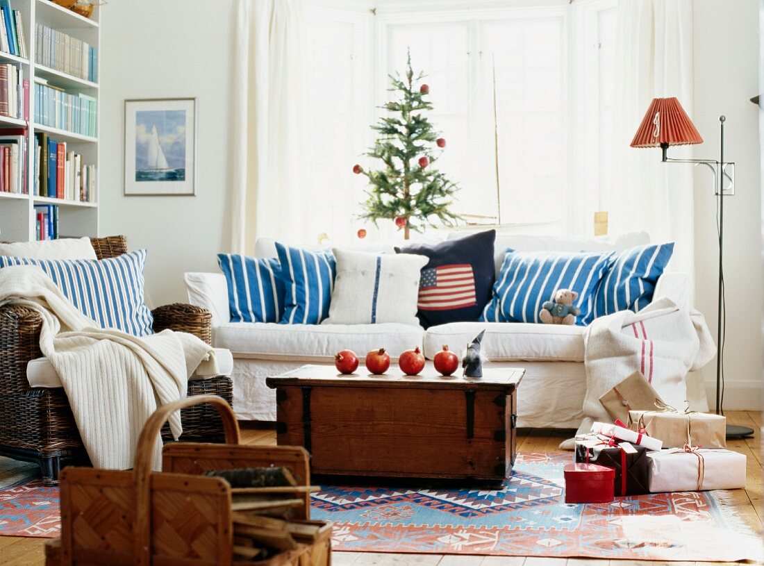 Pomegranates on wooden trunk in front of white sofa with blue and white striped scatter cushions below window in living room decorated for Christmas