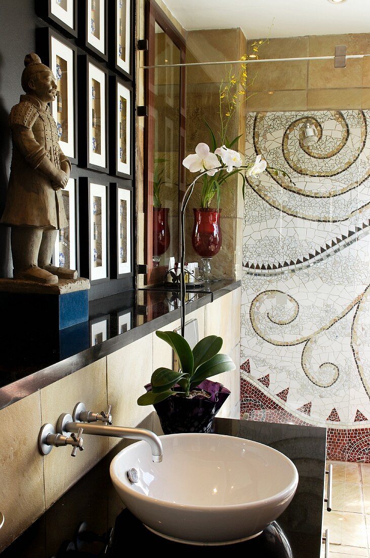 weiße Waschschüssel auf schwarzer Steinplatte und asiatische Figur auf Ablage neben Duschbereich mit Mosaikbild an Wand