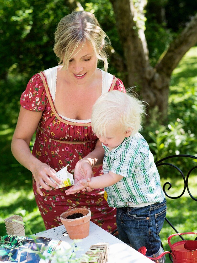 Mutter und Kleinkind am Gartentisch beim Aussäen von Tütensamen in Tontopf