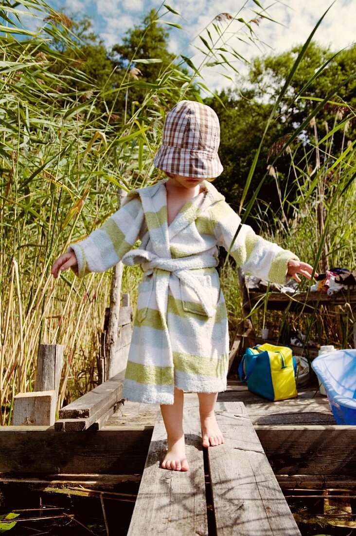 Kleines Kind mit Bademantel und Sonnenhut an Schilfufer auf Holzsteg balancierend