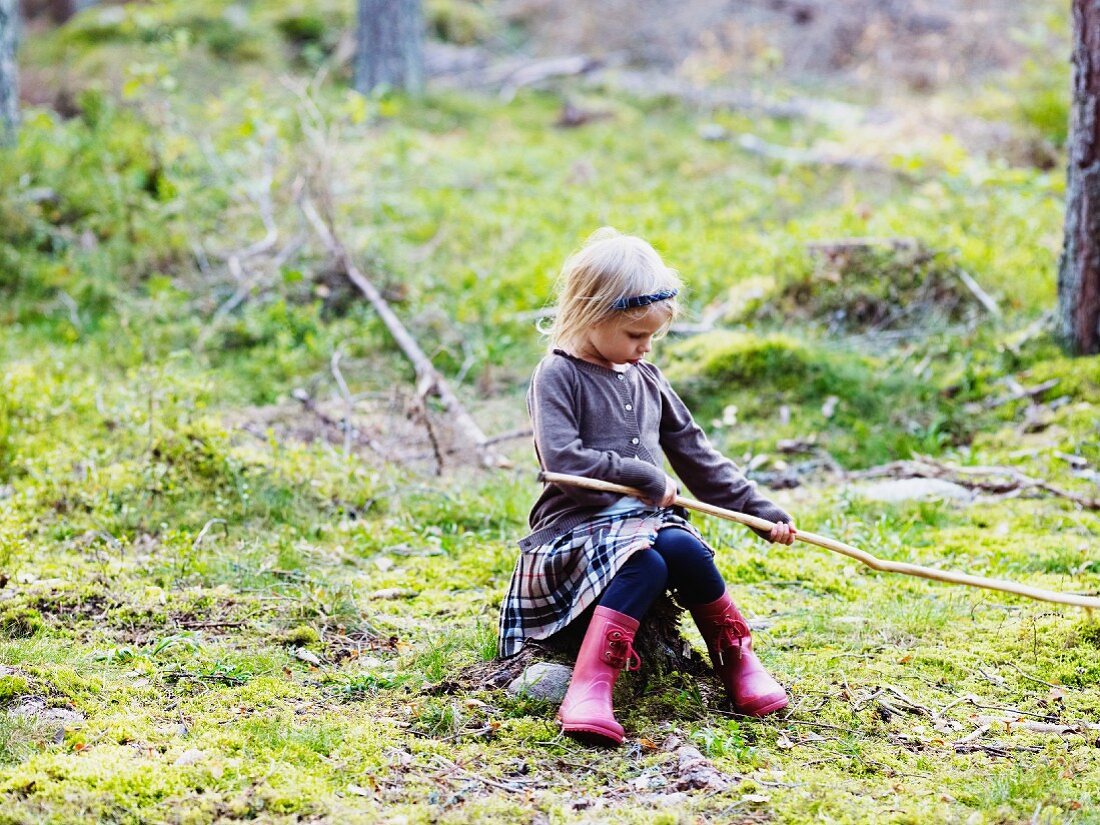Mädchen sitzt auf Baumstumpf einer Waldlichtung und spielt mit einem Stock