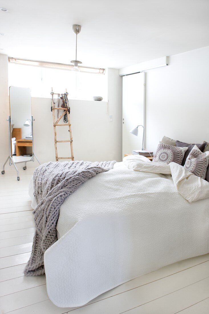Double bed with pale grey, knitted blanket in Scandinavian bedroom with white wooden floor; decorative ladder and full-length mirror in background
