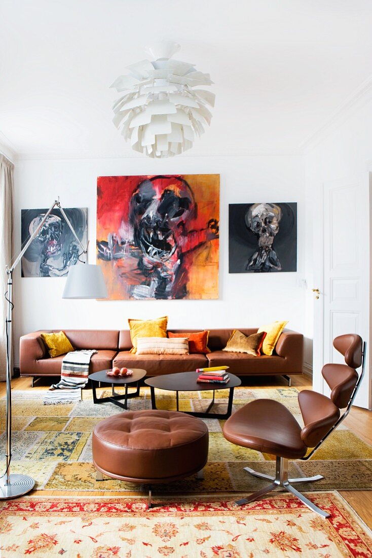 Klassiker Bürosessel mit braunem Lederbezug und passendem Fussschemel vor Loungebereich mit schwarzen Couchtischen und braunem Ledersofa, an Wand expressive Gemälde
