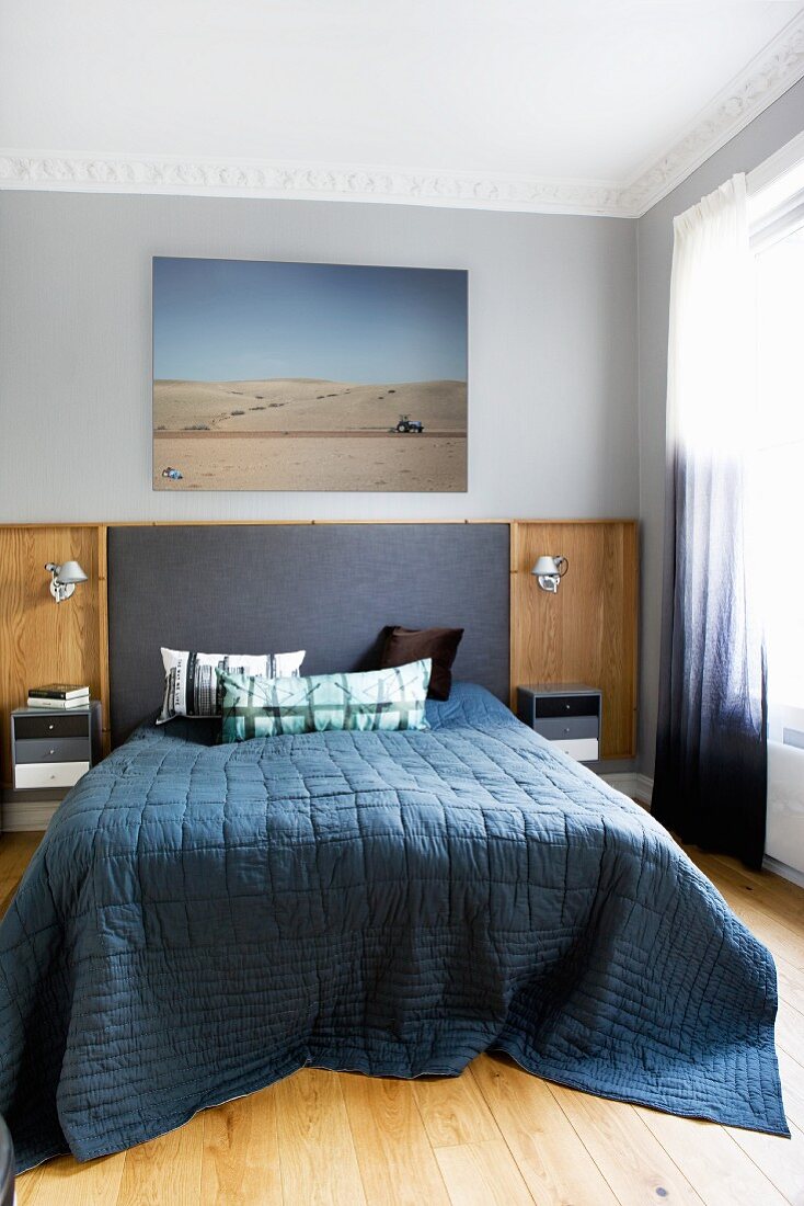 Doppelbett mit dunkelblauem Plaid vor Wand mit teilweise gepolstertem Paneel, darüber Foto mit Wüstenmotiv in traditionellem Schlafzimmer