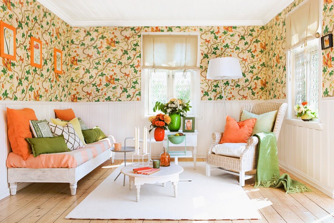 Lounge area with floral wallpaper above dado rail, white wall panelling below and orange and green accessories combined with white furniture