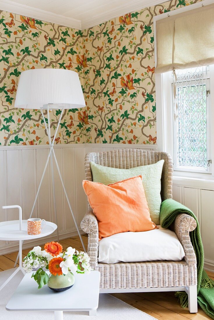 Helle Zone mit Korbstuhl, Stehlampe und Beistelltischen vor brüstungshoher Wandvertäfelung, getoppt von floral gemusterter Tapete; Accessoires und Blumen in Orange und Grün