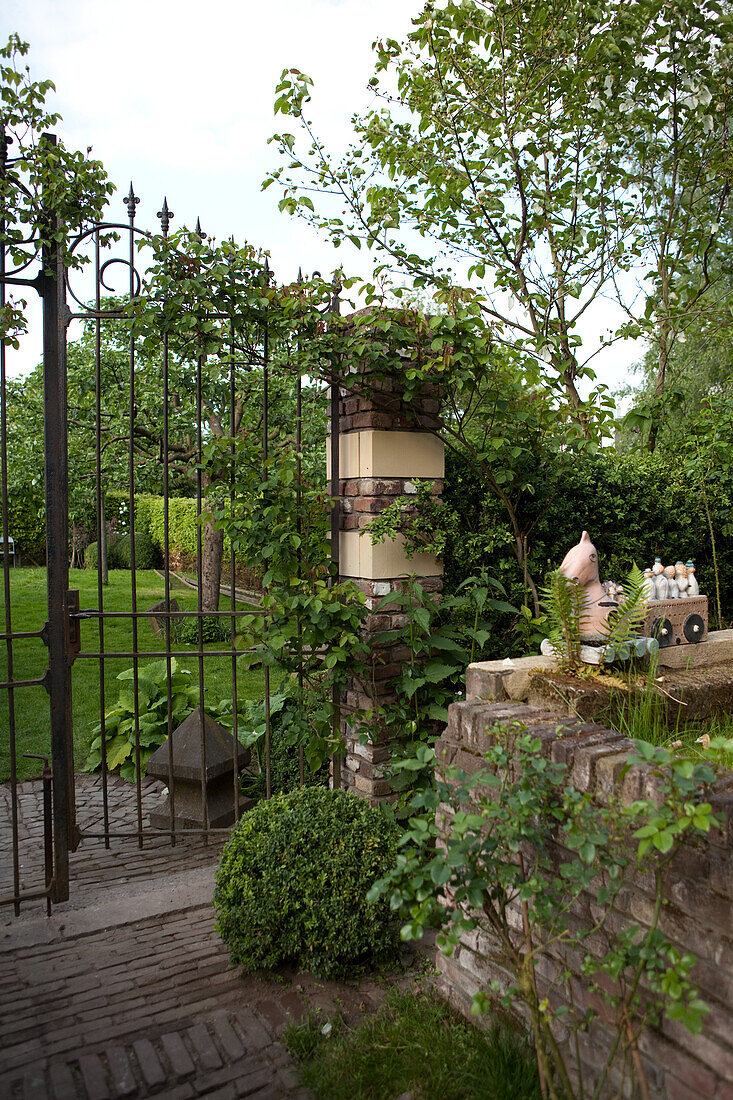 Backsteinmäuerchen und geöffnetes schmiedeeisernes Gartentor in eingewachsenem idyllischem Garten