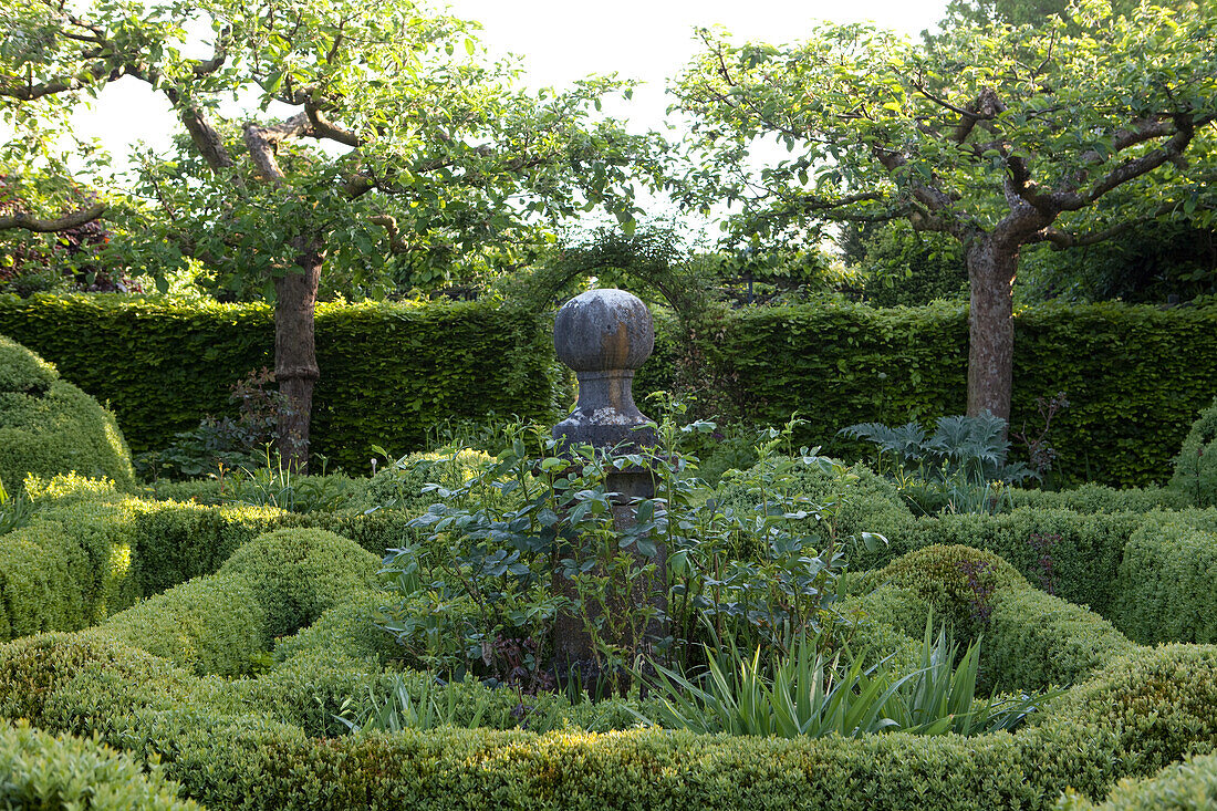 Eingewachsene Skulptur inmitten von geschnittenen Buchsbaumhecken im sonnigen Garten