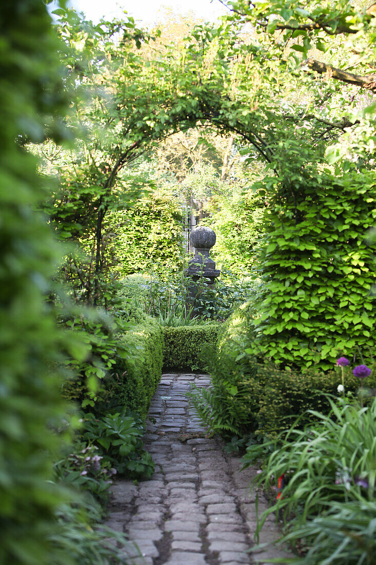 Romantischer Blick auf Steinskulptur durch einen berankten Rundbogen in üppig eingewachsenem Garten
