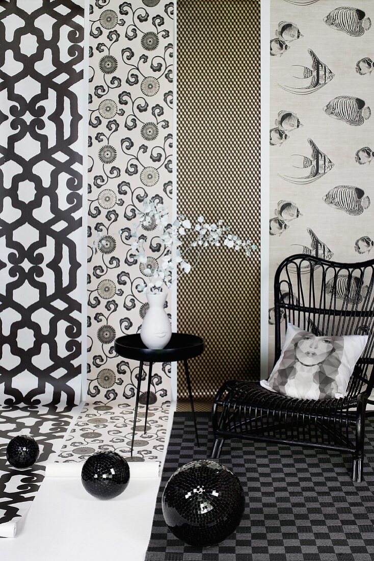 Black disco balls, side table, wicker chair and scatter cushion with print portrait in front of lengths of patterned wallpaper