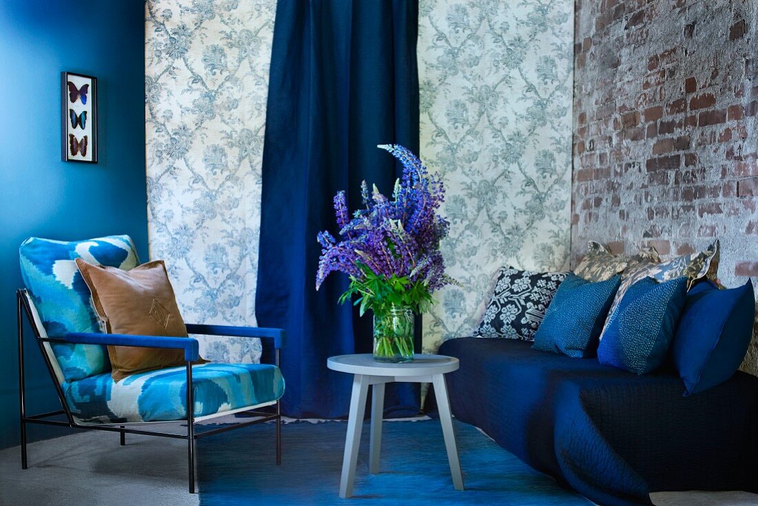 Blau getönte Wand, gemusterte Tapete und Backsteinmauer als Hintergrund für blauen Sitzbereich mit Sofa, Sessel und Lupinenstrauss auf Beistelltisch